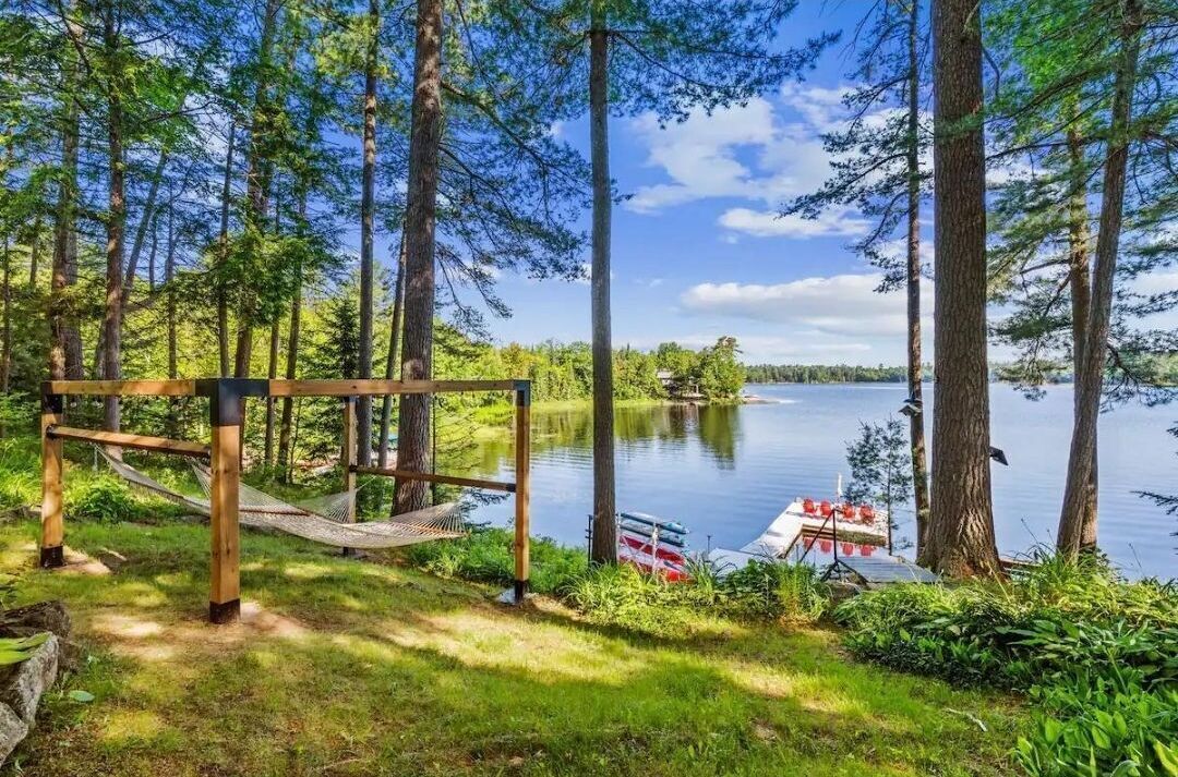 A hammock and wooden structure in a forested area with a lake and a boat docked nearby.