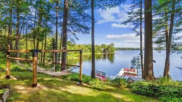 A hammock and wooden structure in a forested area with a lake and a boat docked nearby.