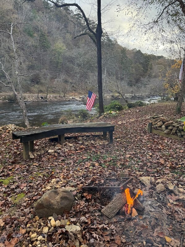 A riverside park with a wooden bench, a fire pit, and an American flag.