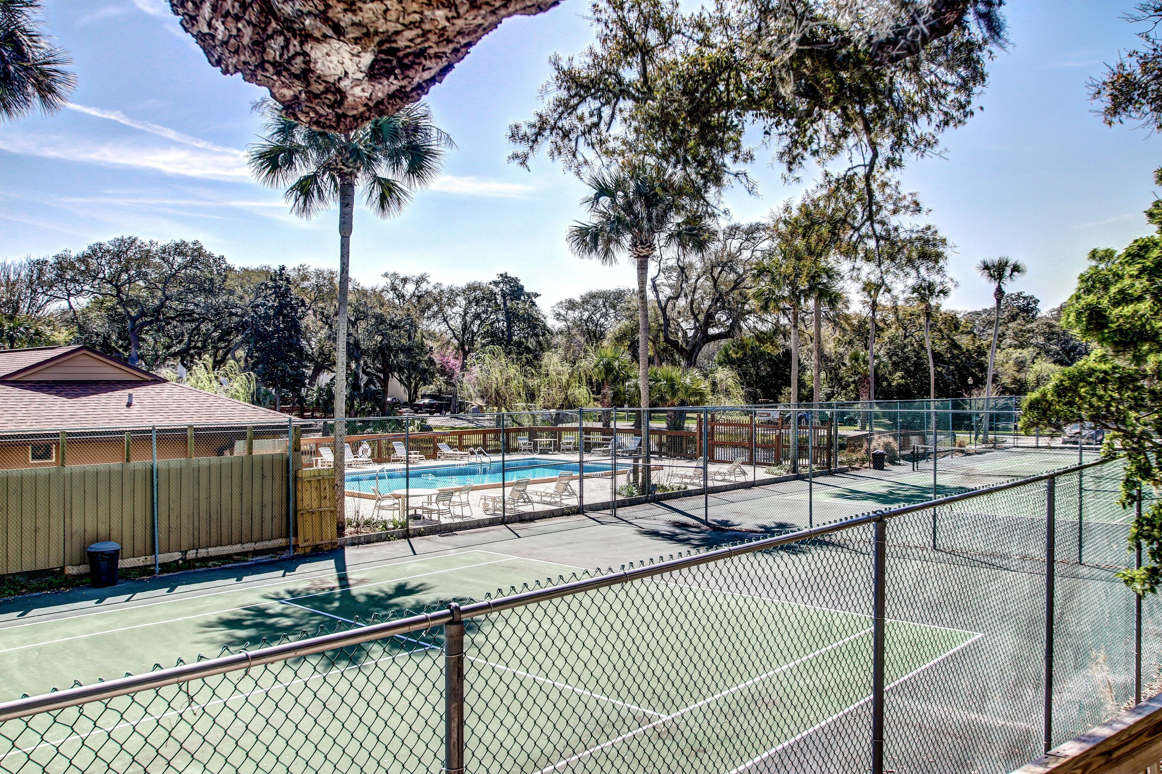 A tennis court with a chain-link fence, a pool area with lounge chairs, and a building with a brown roof.
