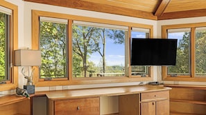 A room with a wooden desk, a flat-screen TV mounted on the wall, and large windows offering a view of trees and a distant landscape.