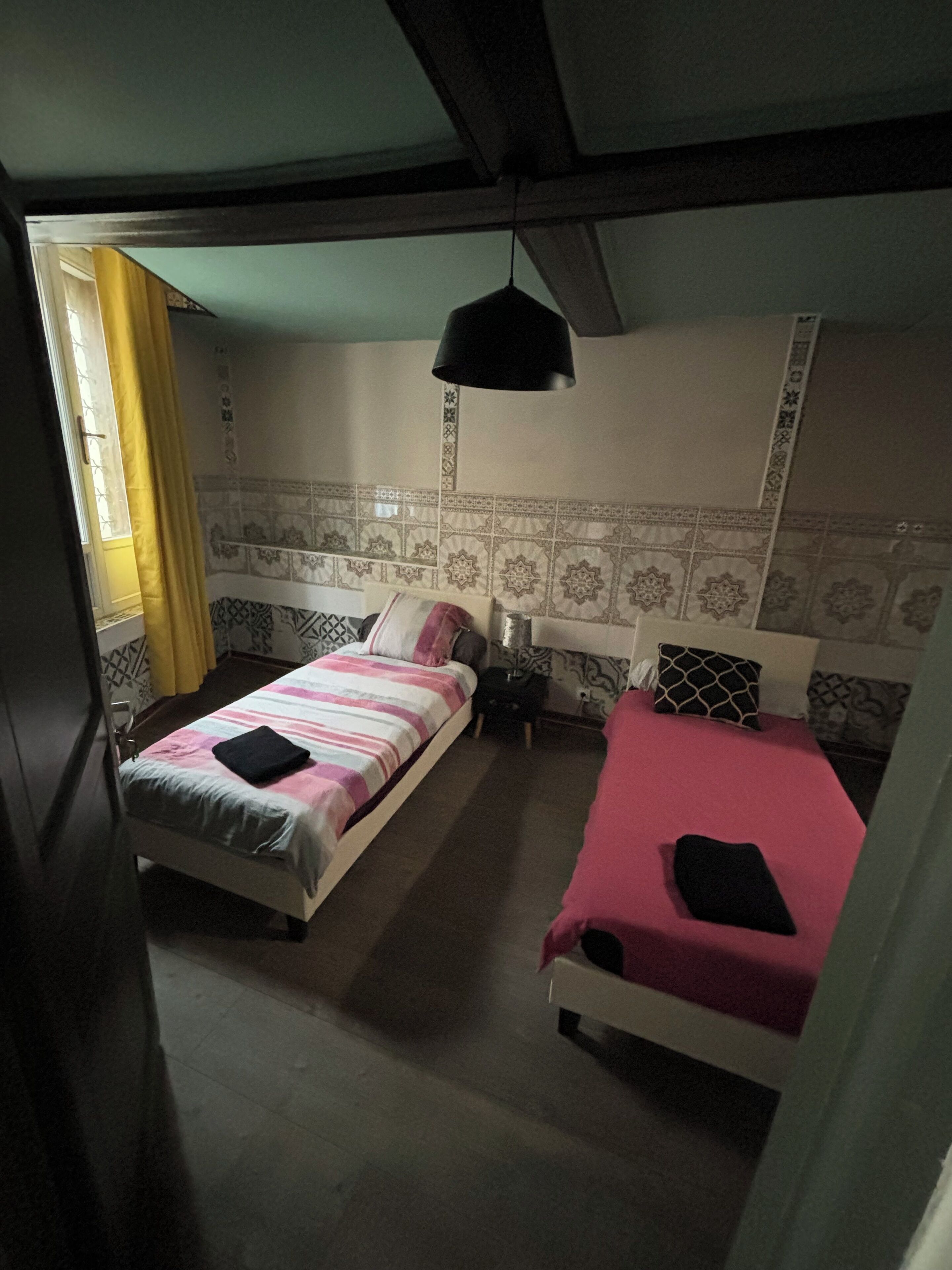 Een kamer met twee bedden, één met een gestreept dekbed en de ander met een effen kleur. Er staat een klein nachtkastje met een lamp erop, en er is een raam met gordijnen.