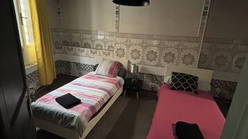 Een kamer met twee bedden, één met een gestreept dekbed en de ander met een effen kleur. Er staat een klein nachtkastje met een lamp erop, en er is een raam met gordijnen.