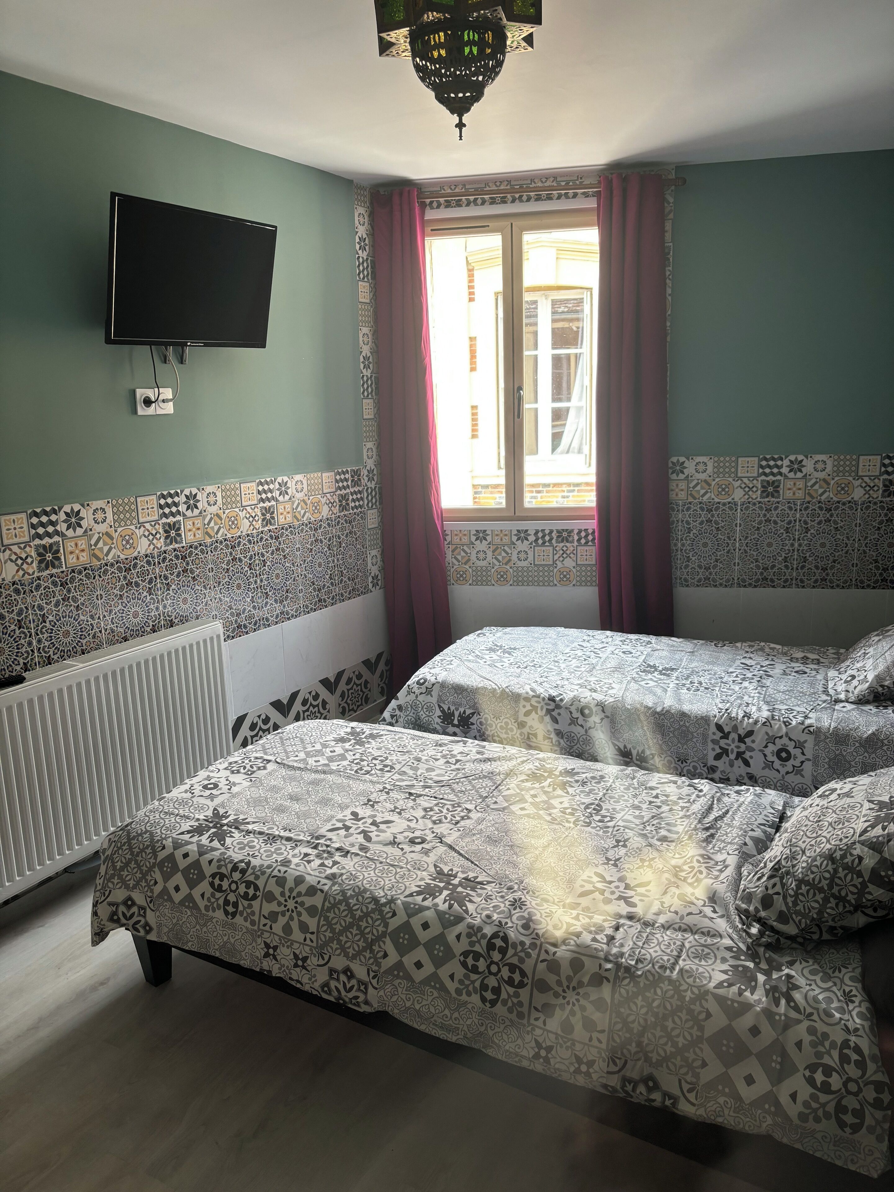 A room with two beds, a TV mounted on the wall, patterned tiles on the walls, a window with curtains, and a radiator.