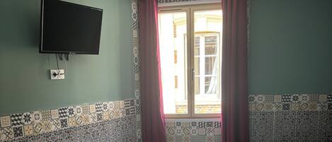 A room with two beds, a TV mounted on the wall, patterned tiles on the walls, a window with curtains, and a radiator.