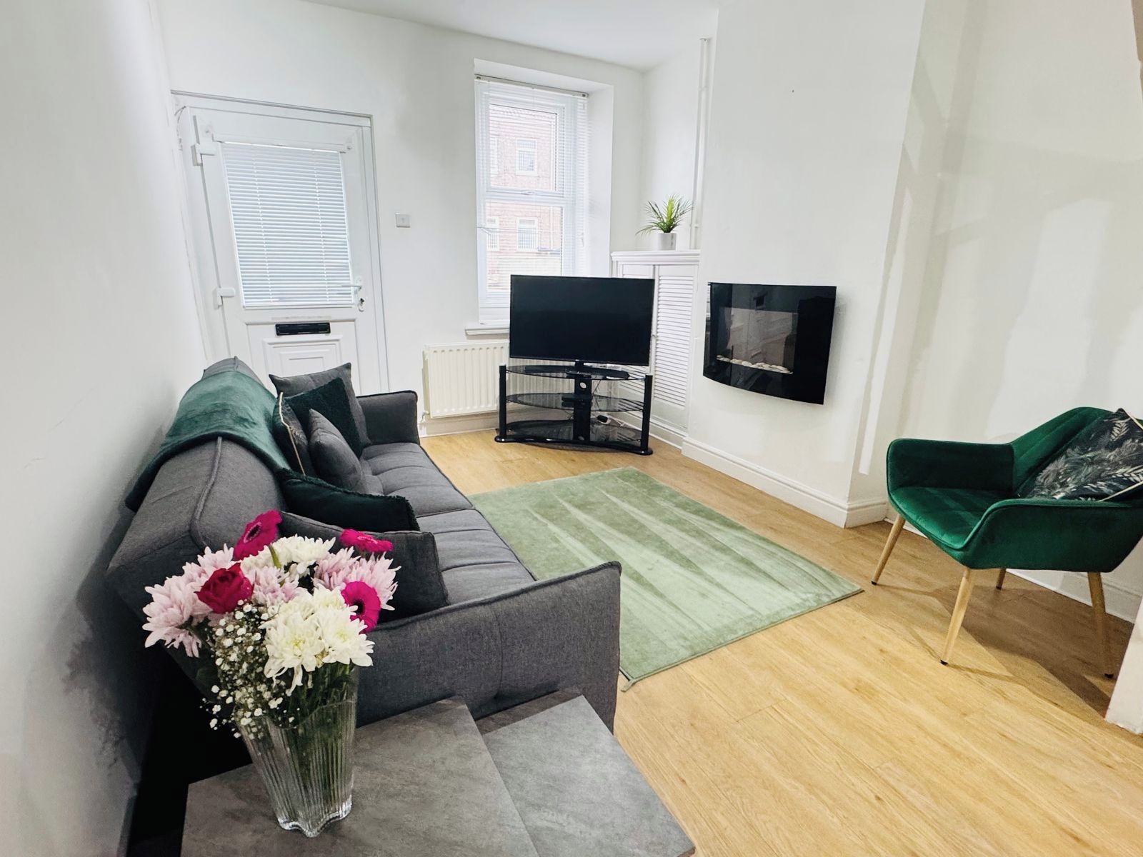 Ein modernes Wohnzimmer mit einem grauen Sofa, einem grünen Sessel, einem Flachbildfernseher und einem kleinen Tisch mit einer Blumenvase.