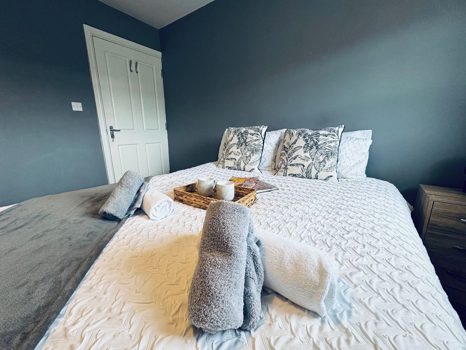 A neatly made bed with a white quilt, two pillows with a leaf pattern, and a tray with a mug and a book.
