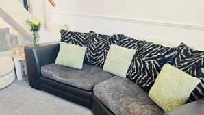 A black leather sofa with grey and green cushions, a small white side table with a glass, and a vase with flowers.
