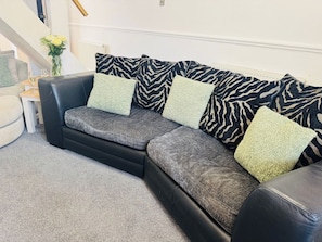A black leather sofa with grey and green cushions, a small white side table with a glass, and a vase with flowers.