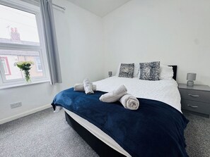 A modern bedroom with a large bed, two bedside tables, a window with curtains, and a vase with flowers on the windowsill. - Bolton Terrace | 2 Bedrooms | Sleeps 4 | Workington (Workington)