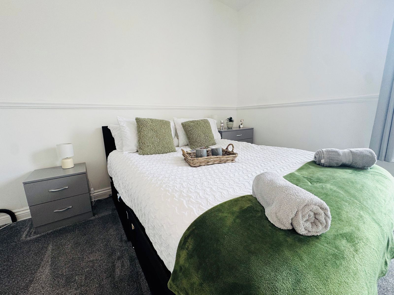 A neatly made bed with a green blanket and rolled towels, a bedside table with a lamp, and a small basket with items on it.