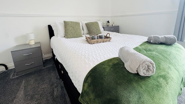 A neatly made bed with a green blanket and rolled towels, a bedside table with a lamp, and a small basket with items on it.