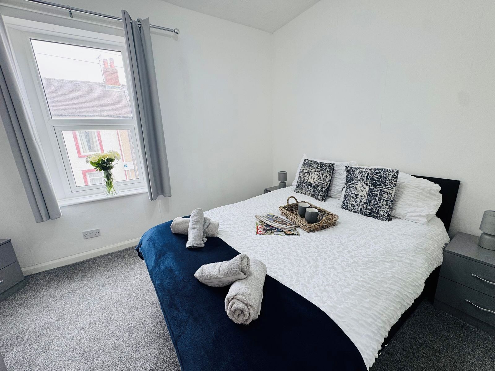 Ein ordentlich bezogenes Bett mit weißer Daunendecke, zwei Kissen und einer blauen Decke. Auf dem Bett steht ein Korbwäscher mit zwei Tassen und einer Zeitschrift. Auf dem Fensterbrett steht eine Vase mit Blumen.