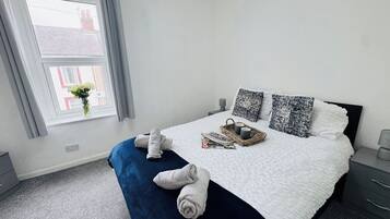 A neatly made bed with a white duvet, two pillows, and a blue blanket. There is a wicker basket with two mugs and a magazine on the bed. A vase with flowers is on the windowsill.