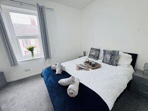 A neatly made bed with a white duvet, two pillows, and a blue blanket. There is a wicker basket with two mugs and a magazine on the bed. A vase with flowers is on the windowsill.