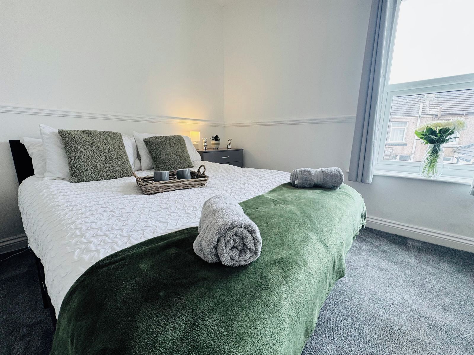 A hotel room with a bed, a green throw, a tray with two mugs, and a window with a view of buildings.