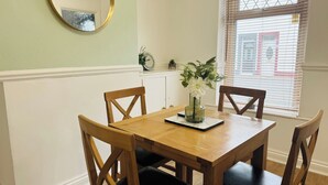A dining area with a wooden table and chairs, a vase with flowers, a clock, and a window with blinds.