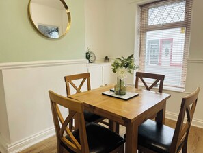 A dining area with a wooden table and chairs, a vase with flowers, a clock, and a window with blinds.