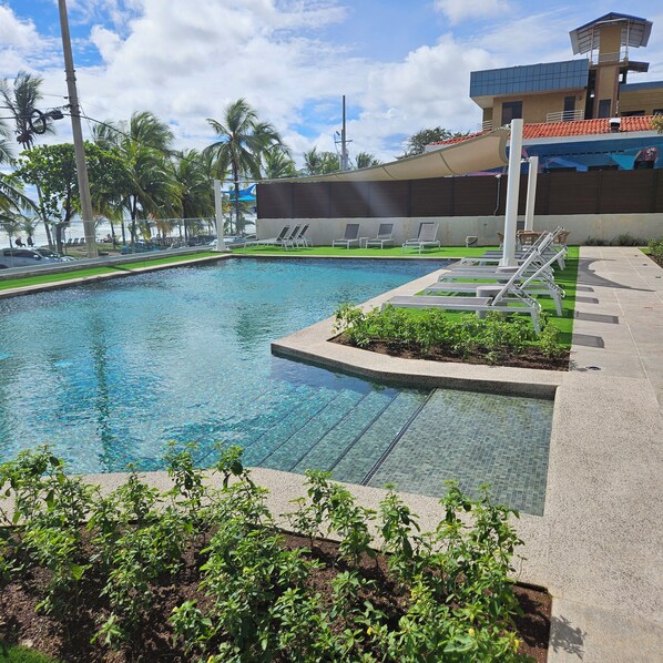 Una piscina cristalina con sillas de descanso y vistas a palmeras y un edificio al fondo.