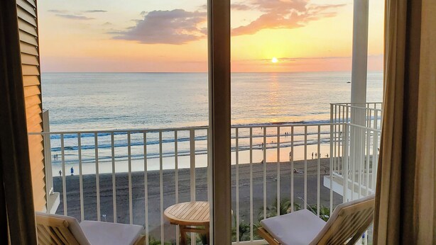 Un balcón con dos sillones de descanso y una pequeña mesa, con vista a una playa donde hay gente y un atardecer.