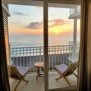 Un balcón con dos sillones de descanso y una pequeña mesa, con vista a una playa donde hay gente y un atardecer.