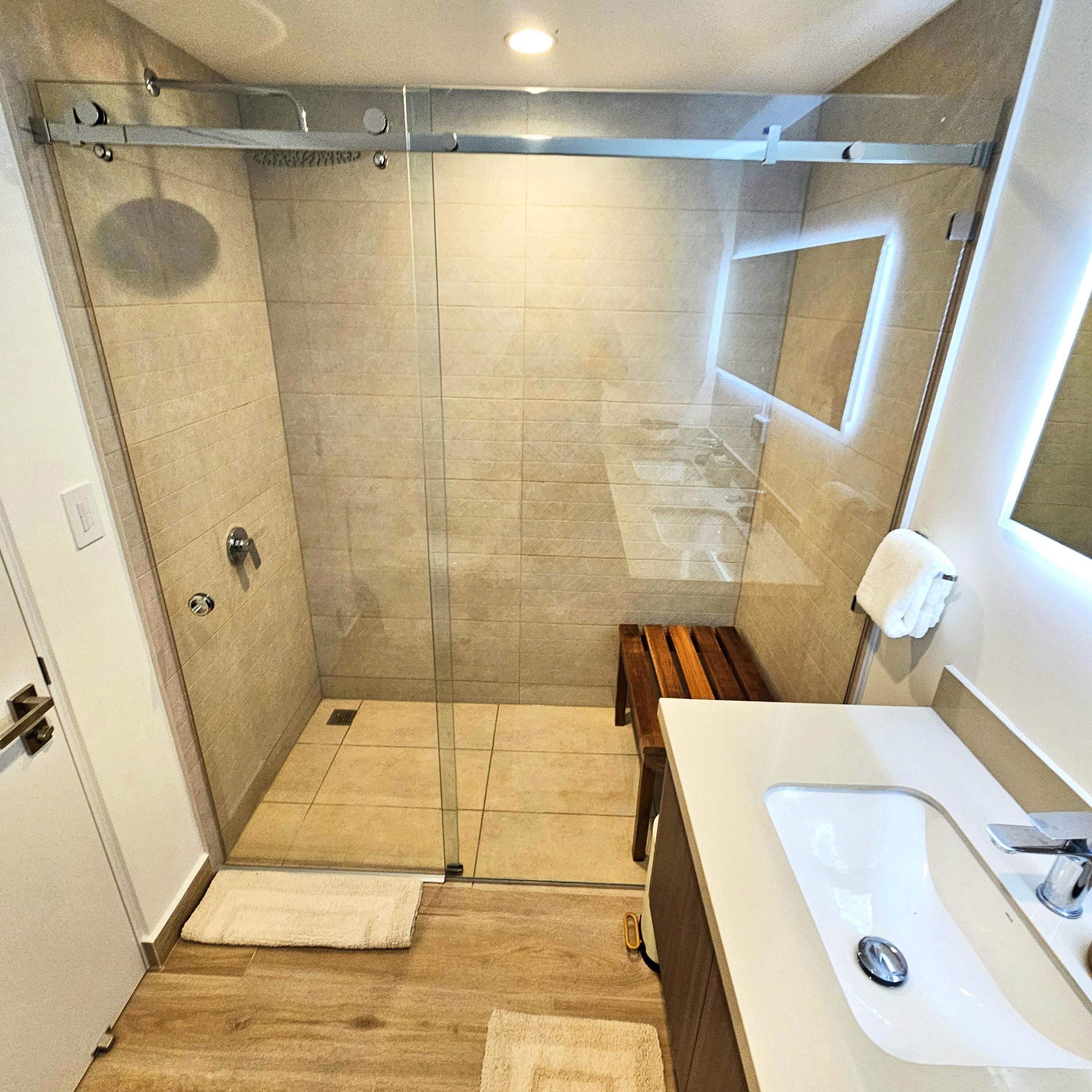 A modern bathroom with a glass shower enclosure, a wooden bench, and a white sink.