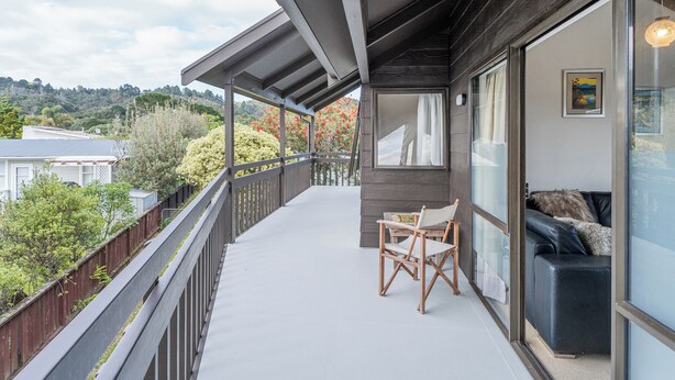 A balcony with a wooden chair and a couch, a view of trees and houses, and a painting on the wall.