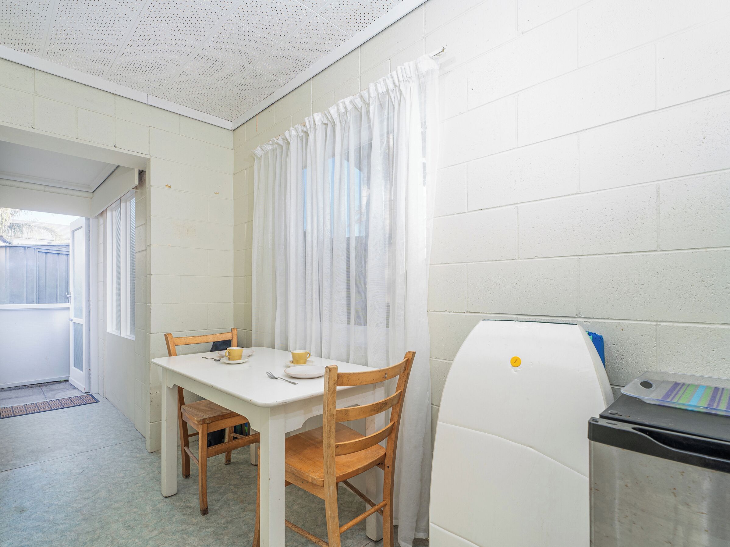 A small kitchen with a white refrigerator, a wooden table with chairs, and a window with white curtains.