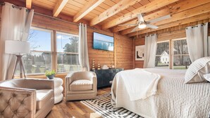 Un dormitorio de cabaña de madera con cama, una silla, un escritorio, un televisor y una ventana con cortinas.