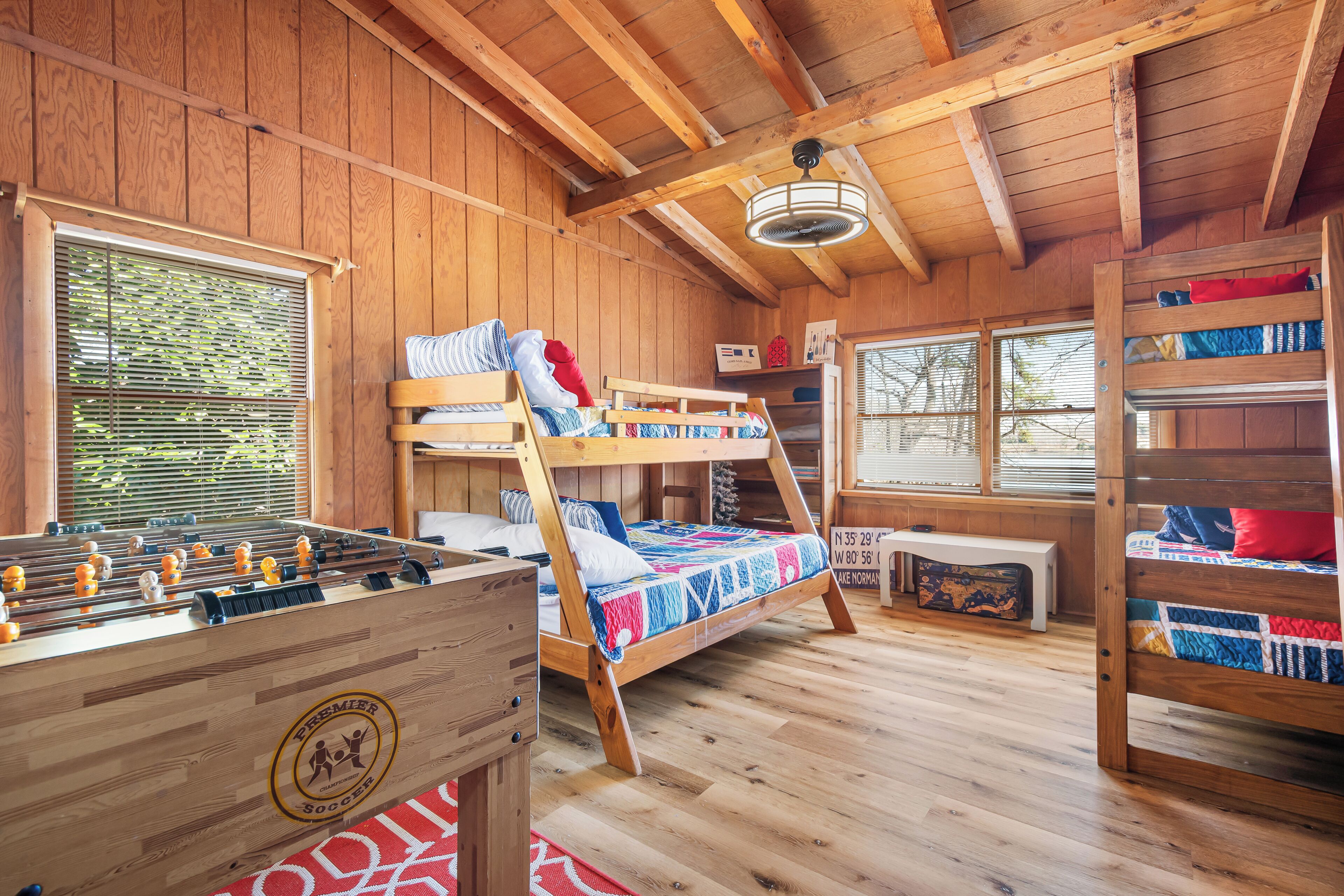 A wooden-floored room with bunk beds, a foosball table, and a window with blinds.