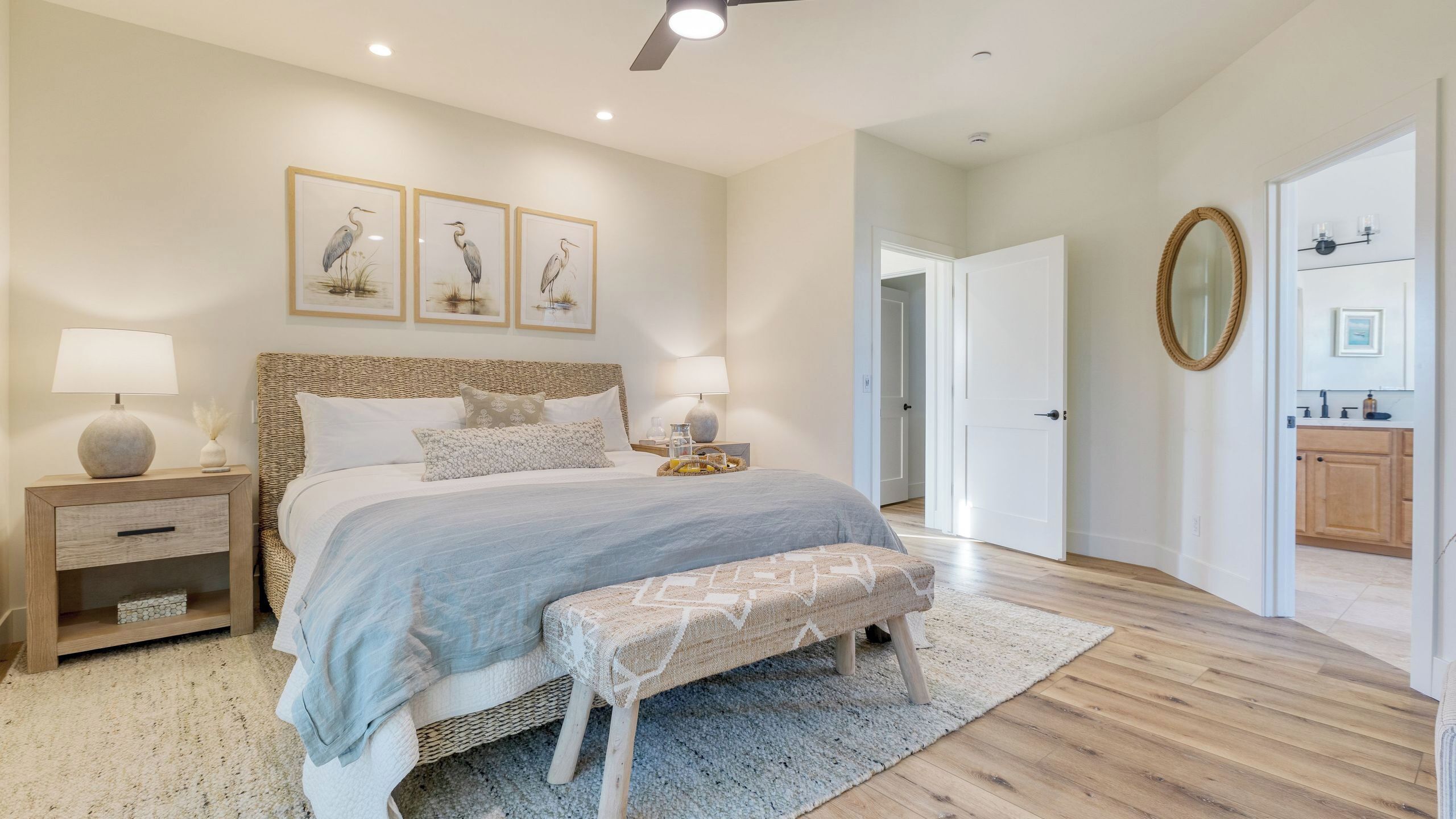 Une chambre à coucher comprenant un lit, des tables de chevet, un banc, un ventilateur de plafond, des œuvres d’art encadrées, un miroir et une porte donnant sur une salle de bain.