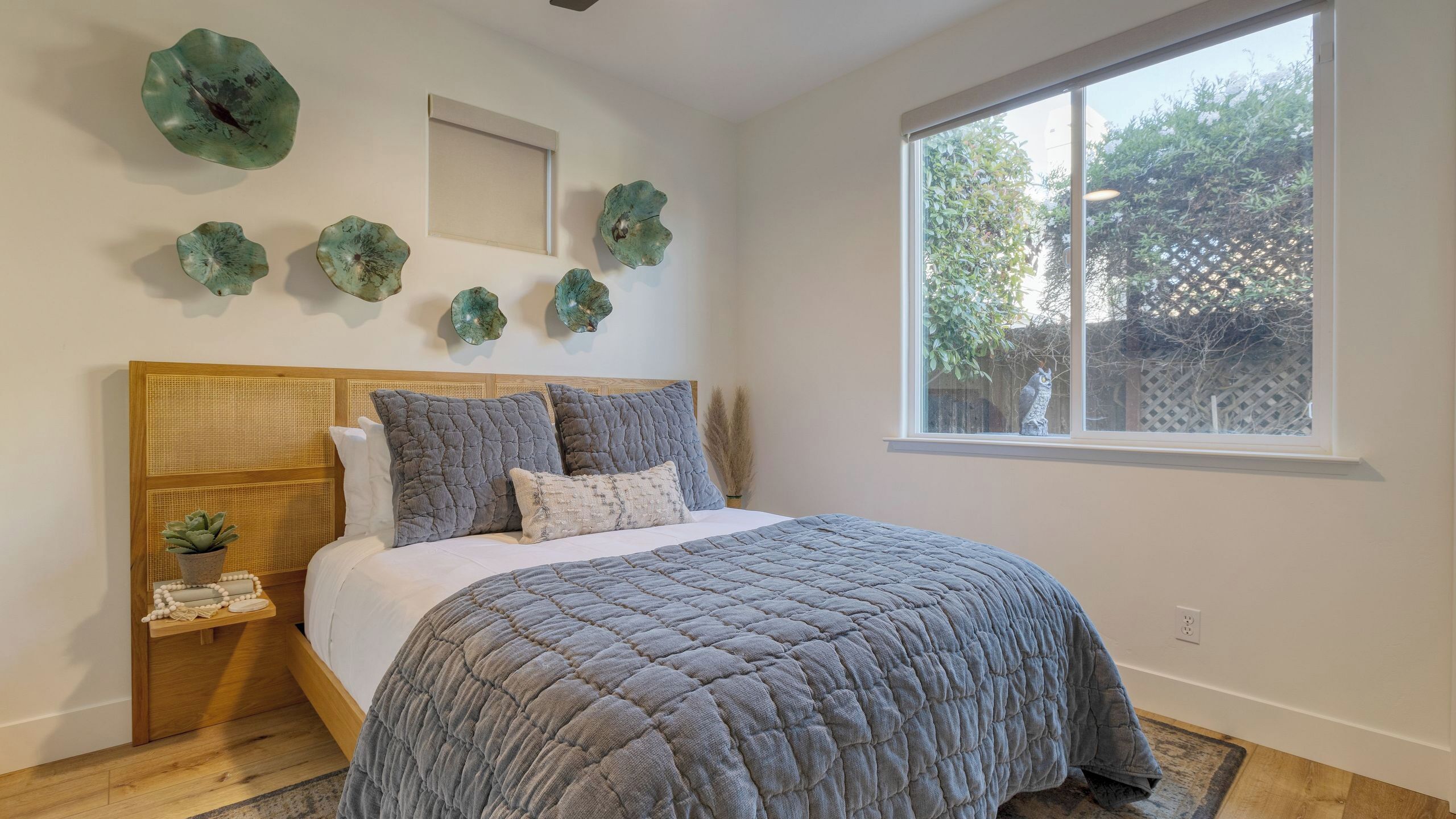 Une chambre à coucher avec une tête de lit en bois, un grand lit avec une literie grise, une fenêtre donnant sur un paysage verdoyant et des éléments décoratifs fixés au mur.