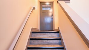 Un escalier avec des marches en bois et une rampe métallique, menant à une porte dotée d’une fenêtre et d’un luminaire placé au-dessus.