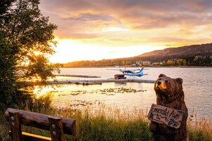 Un banc en bois et une statue d’ours tenant une pancarte, le tout situé au bord d’un lac, avec un coucher de soleil en arrière-plan.