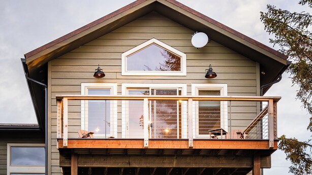 Une cabane en rondins à deux étages, dotée d’un balcon et d’une terrasse, avec de nombreuses fenêtres et une antenne parabolique.