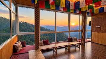A room with large windows offering a mountain view, a wooden bench, and colorful prayer flags.