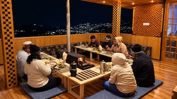 A group of people seated around a wooden table with a wood-burning stove, dining on a terrace with a city view at night.