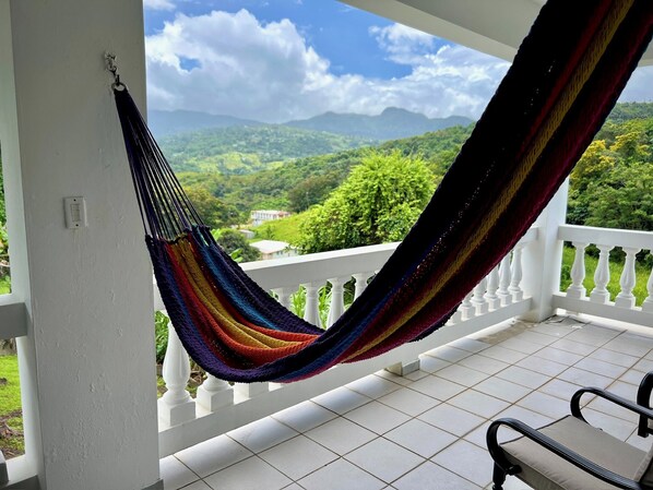 A hammock on a balcony with a view of green hills and a house in the distance.
