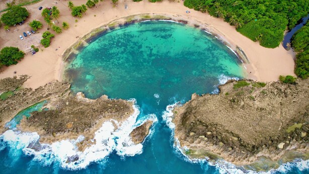 An aerial view of a beach with clear turquoise waters, a sandy shore, and rocky outcrops.