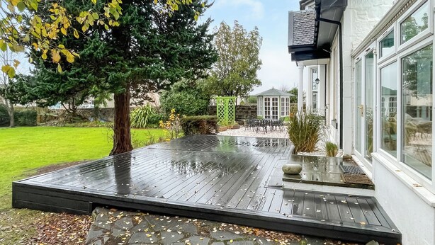 Une terrasse en bois avec un revêtement de sol en pierre, un arbre et un bâtiment blanc doté de grandes fenêtres.