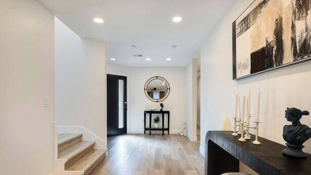 A hallway with wooden flooring, a staircase, a black console table with candles, a bust sculpture, and a round mirror on the wall.