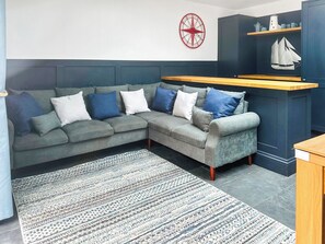 Ein graues Ecksofa mit blau-weißen Kissen, ein gemustertes Teppich, eine rote Wanduhr und eine dunkelblaue Schrank mit Regalen darüber.