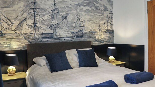 Ein Bett mit weißer Bettwäsche, zwei dunkelblauen Kissen und zwei aufgerollten blauen Handtüchern. Zwei Nachttische mit weißen Lampenschirmen stehen bereit. An der Wand hinter dem Bett befindet sich ein großes, detailliertes Wandbild mit Segelschiffen vor einer strukturierten Hintergrundkulisse.