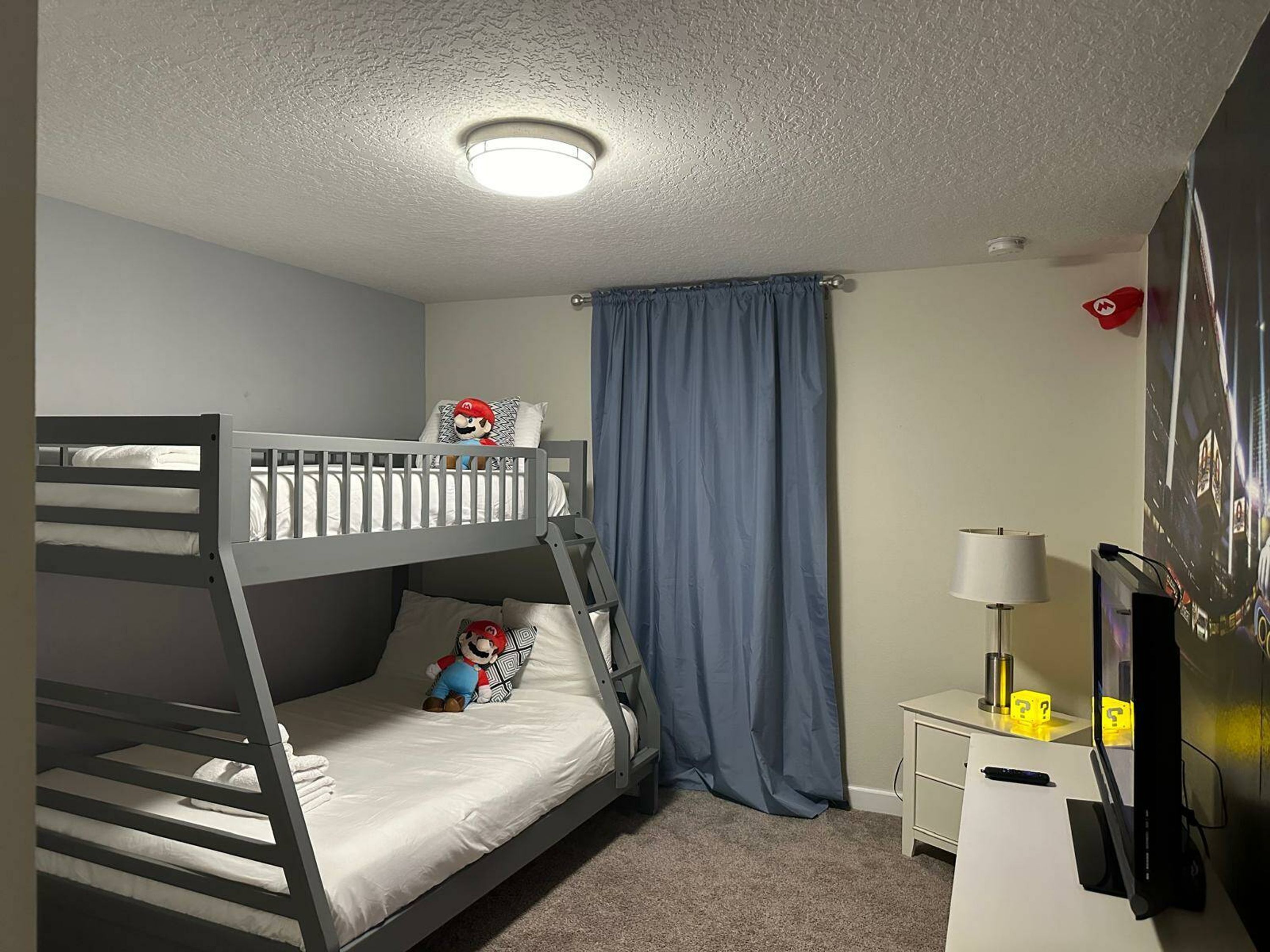 Un lit superposé avec un oreiller à l’effigie de Mario, un bureau avec une lampe et un téléviseur posé sur un meuble.