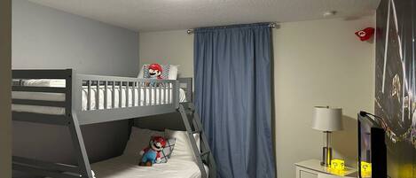 Un lit superposé avec un oreiller à l’effigie de Mario, un bureau avec une lampe et un téléviseur posé sur un meuble.
