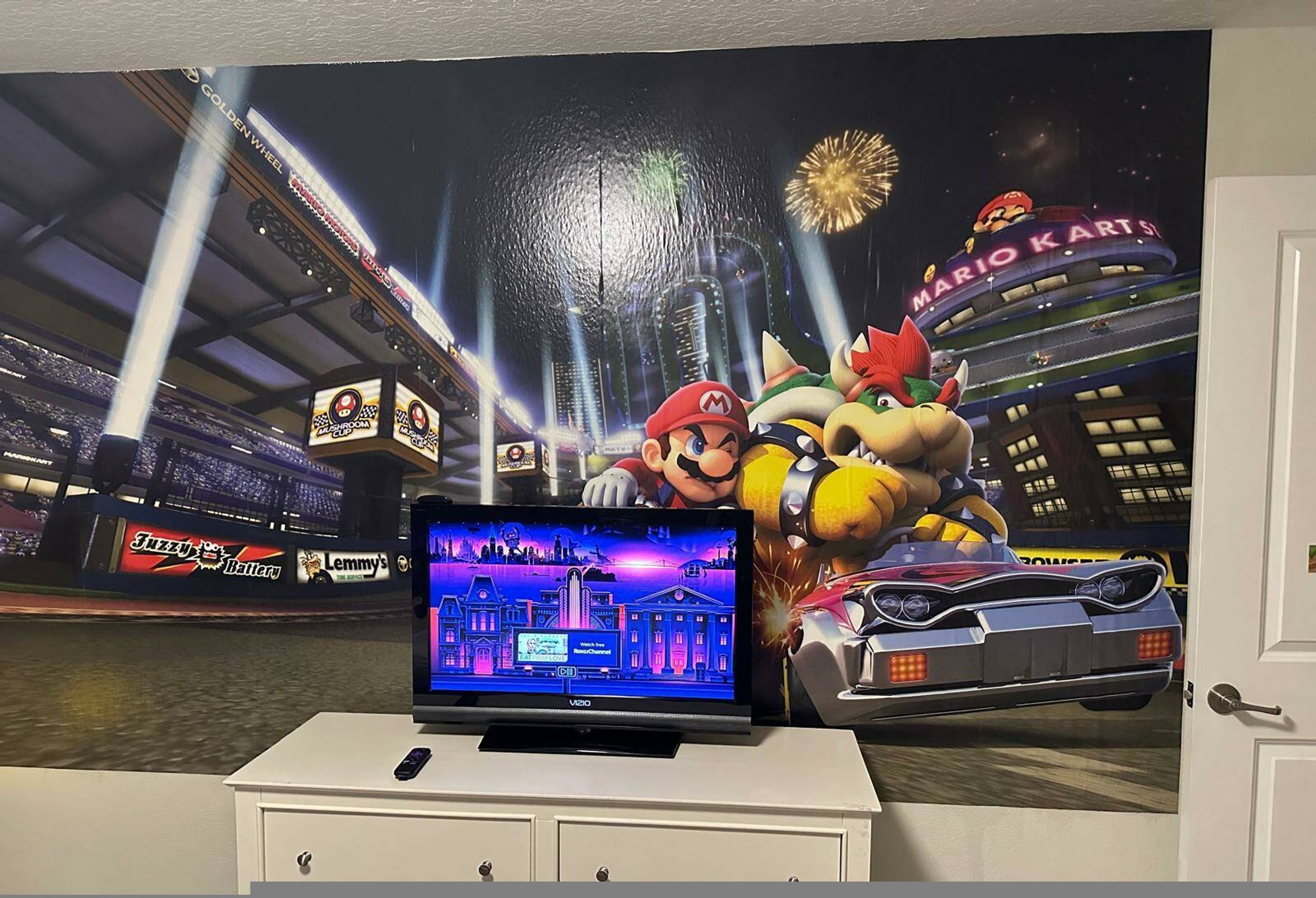 Ein Fernseher, der ein Mario Kart Spiel zeigt, eine Wand mit Mario Kart Motiven und ein Modellrennwagen.