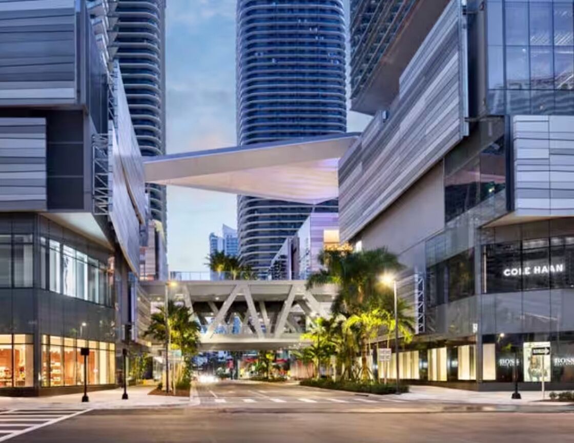 A modern urban street with high-rise buildings, a pedestrian bridge, and storefronts including a Cole Haan store.
