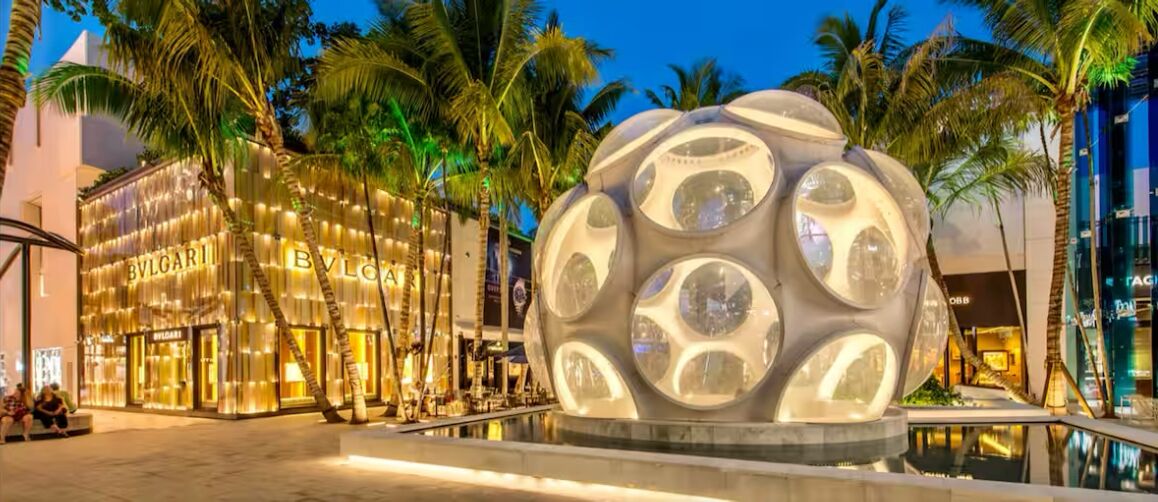 A luxury hotel with a distinctive spherical glass structure in the foreground, a well-lit storefront with the name 'BVLGARI' on the building, and palm trees in the background.