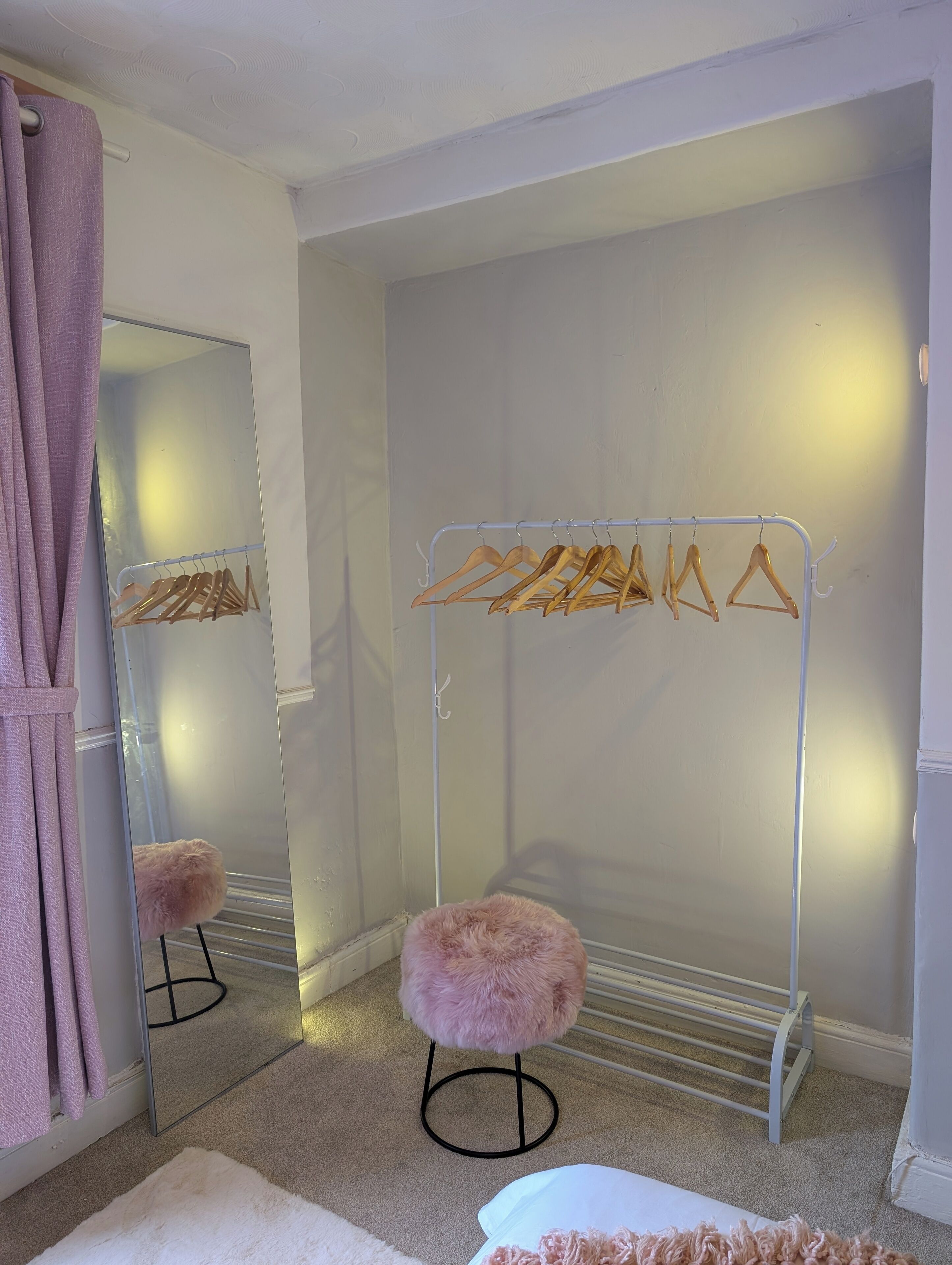 A room with a mirror, a metal drying rack with wooden hangers, a fluffy pink stool, and a white rug.
