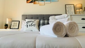 Cama bem arrumada com toalhas dobradas, cabeceira cinza e parede com três quadros retratando paisagens tropicais.